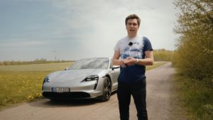 Mann steht vor grauem Porsche Taycan mit Feld und Windrad im Hintergrund